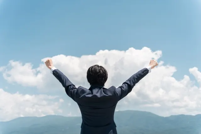 スーツ姿の男性が青空と山並みに向かって両手を挙げている後ろ姿