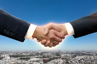 青空と街並みを背景に、スーツ姿の2人が握手している手元
